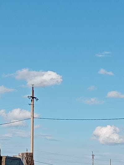 The image shows a clear blue sky with scattered white clouds. A tall, slender utility pole with wires running horizontally across it is visible in the center of the frame. At the base of the pole, a partially visible structure that appears to be a building or a concrete platform can be seen. In the background, a few more clouds are present, and a thin, dark line, likely another wire, stretches across the lower portion of the image. The scene appears to be outdoors and could be located in the city of Oktyabrskoye, Kazakhstan, given the context.