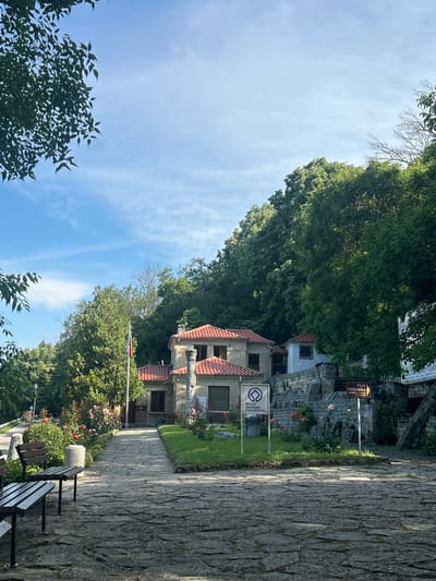 The image shows a stone pathway leading to a complex of stone buildings with red tiled roofs, situated on a hillside covered in dense green trees. In the foreground, there are two wooden benches facing the pathway. To the left of the pathway, there is a patch of grass with red roses and a cylindrical stone object. To the right of the pathway, there is a green lawn with rose bushes and a stone wall. A sign with the text "World Heritage Convention" is visible on the left side of the pathway. A yellow directional sign with text in Cyrillic is posted on the right side of the pathway. A Bulgarian flag is planted near the buildings. The sky is blue with scattered white clouds. The scene appears to be an outdoor tourist attraction or historical site.