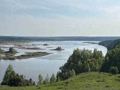 The image depicts a broad, calm lake landscape near Serga, Russia. Several small, mostly flat islands are visible in the lake, some with light vegetation along their edges. The shoreline of the lake is heavily forested. In the foreground, a grassy hillside is partially covered with trees and shrubs. The sky is overcast with a few sparse clouds.