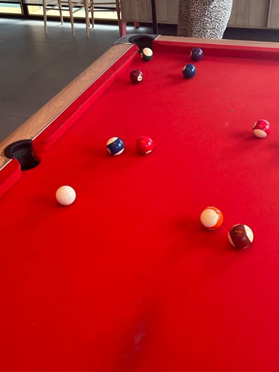 This image depicts a red-felted pool table in an indoor setting within Garston, United Kingdom. The table features wooden rails with metallic corner accents; two pockets are visible, one in the foreground left and one in the mid-right. Ten pool balls are distributed across the red surface: one white cue ball, two solid blue balls, one solid red ball, one solid dark purple ball (numbered '7'), one solid dark green ball (numbered '6') partially submerged in the top right pocket, one striped red and white ball, one striped orange and white ball, and one striped dark brown and white ball. The background reveals a dark grey tiled floor, light-colored wooden chairs, and a wooden cabinet. A leg clothed in leopard print fabric is partially visible behind the table, indicating the presence of an individual.