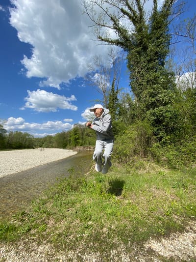 The image captures a bright, sunny outdoor scene in Beregovoe, Russia, featuring a young person by a shallow river or stream. A male-presenting individual, dressed in a grey hooded tracksuit, a white baseball cap, and white Crocs, stands on the grassy bank. They are looking downwards, possibly at something in their hands or observing the ground/water.
The natural setting includes a narrow, clear body of water with a prominent light-colored, gravelly riverbed that extends along the right side of the frame. Adjacent to the water is a wide bank composed of similar light-colored gravel. The banks and background are densely covered with vibrant green grass, various bushes, and trees, some with full foliage and others with bare branches, suggesting either early spring or late autumn with lingering greenery.
The sky in the upper left is a vivid blue, dotted with numerous fluffy white cumulus clouds, indicating pleasant and clear daytime weather. The overall atmosphere of the scene is tranquil and natural. No visible text is present in the image.