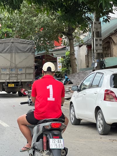 In Hà Giang 2 Ward, Vietnam, an individual wearing a red polo shirt with a white number "1" emblem and black shorts is seated on a scooter. They are also wearing a white helmet. The scooter is gray with red accents. The individual's left leg is visible, and they are wearing a black flip-flop. The scooter's license plate reads "22-AA 915.69". In the background, a large gray truck is parked, with the partial license plate "23C 050.00" visible. A white car is parked to the right of the scooter. Trees and utility poles with multiple meters are also present in the environment. The ground is paved.