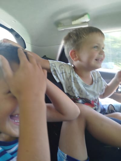 Two young boys are in the back seat of a vehicle. The boy on the right is looking to the right and smiling, with his mouth open. He is wearing a light-colored t-shirt with a graphic print. The boy on the left has his hands covering his face and appears to be laughing. He is wearing a blue and white striped shirt. The interior of the vehicle is visible, including the headliner and a window. The background outside the window is blurred, suggesting motion.