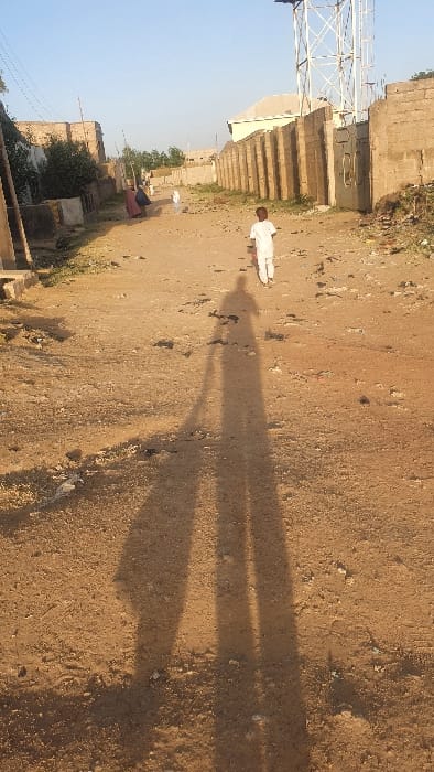 The image displays an unpaved dirt path in Danbare, Nigeria, illuminated by a low sun casting long shadows forward onto the ground, indicative of morning or late afternoon. In the foreground, two elongated human shadows are prominent on the reddish-brown earth, which is scattered with small rocks and debris. A young child, dressed in white attire, walks away from the viewer in the mid-ground, positioned centrally on the path. Further back on the left side of the path, an adult figure in dark, possibly maroon, clothing is either seated or crouched near another smaller, lighter-clad individual. The path is bordered on the left by a partially visible structure made of concrete blocks and some green foliage. On the right, a long wall constructed from concrete blocks extends into the background, culminating in a metal gate. Beyond this wall, a light-colored building is visible. A tall, white lattice-style communication tower stands against a clear, light blue sky in the upper right background.