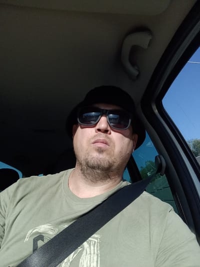 This media file shows a man wearing a dark bucket hat and sunglasses, with a light green t-shirt and a black seatbelt across his chest. He is sitting in the driver's seat of a car, with the interior of the vehicle visible around him. The sky outside is clear and blue, suggesting it is daytime with good weather. The sun is casting shadows, indicating it might be mid-morning or afternoon.
The man appears to be looking forward, possibly driving, and has a neutral expression. The interior of the car shows a grab handle on the roof and the edge of the car door. Through the window, trees and a fence are visible, suggesting a suburban or rural setting. There is no visible text in the image. The overall impression is a candid shot of someone on a drive. The location context of Kunice, Czechia is provided.