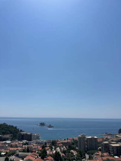 The image depicts a panoramic view of the coastal city of Brežine, Montenegro. The foreground features a dense urban landscape with numerous buildings, characterized by terracotta roofs and varying architectural styles. Some buildings appear to be residential, while others might be hotels or commercial establishments, indicated by their size and construction. Lush green vegetation, likely trees and shrubs, is interspersed among the structures, adding a natural element to the cityscape.
In the middle ground, the city gives way to the Adriatic Sea, a vast expanse of deep blue water. The sea appears calm, with a few small boats visible, one creating a white wake as it moves. In the distance, a small cluster of rocky islands, likely Sveti Stefan, emerges from the water. These islands are covered with greenery and appear to have some structures on them, suggesting a historical or resort area.
The sky above is a clear, vibrant blue, devoid of clouds, indicating a sunny day with excellent visibility. The overall impression is one of a picturesque coastal city bathed in bright sunlight, with a striking contrast between the urban development, the natural landscape, and the expansive sea. The viewpoint suggests the photograph was taken from an elevated position, offering a commanding perspective of the city and its surroundings. There are no visible people in the image.