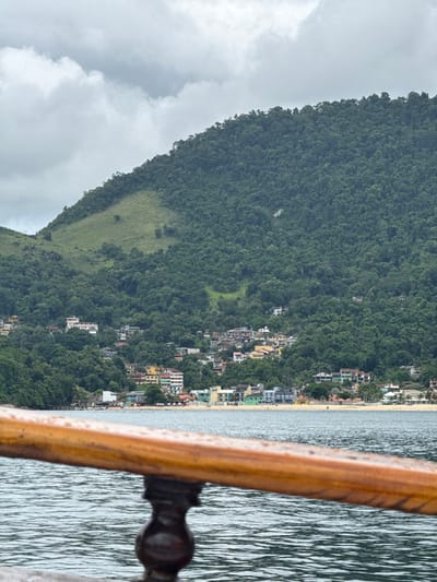 The image depicts a coastal view of the city of Mangaratiba, Brazil, observed from the perspective of a boat on a body of water.
In the immediate foreground, a wet, varnished wooden handrail with visible droplets of water extends horizontally across the lower portion of the frame, partially obscuring the view of the water directly below. A dark, turned wooden baluster supports the handrail.
The midground consists of dark, rippled ocean water under an overcast sky.
In the background, the coastline of Mangaratiba is visible. A light-colored sandy beach stretches horizontally, bordered by a dense line of buildings of varying architectural styles and colors, including yellow, light blue, green, and white facades. Several small, white structures, possibly umbrellas or tents, are present on the beach. Immediately behind these beachfront structures, a steep, heavily forested hill rises dramatically. Numerous residential and commercial buildings are interspersed within the dense green tropical vegetation that covers the lower and mid-slopes of this hill. A distinct patch of lighter green, grassy terrain is visible on the upper left slope of the hill, contrasting with the surrounding dark green forest. The upper section of the hill and the sky are dominated by heavy, uniform grey clouds, indicating an overcast day. No discernible individuals or specific actions are visible.