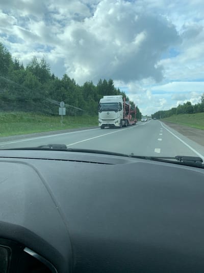The image shows a view from the interior of a vehicle, looking out at a highway. A white car carrier truck is visible on the highway, carrying cars on its trailer. The truck has a distinctive front grille with a pattern of hexagons. Several other vehicles are also on the road, appearing to be cars. The highway is flanked by dense green trees and vegetation on the left and right sides. The sky is partly cloudy with patches of blue sky visible between the clouds. The foreground of the image shows the dashboard of the vehicle, including the windshield wipers. The scene appears to be set on a sunny day, possibly in the summer. The location is identified as Nevolino, Russia.