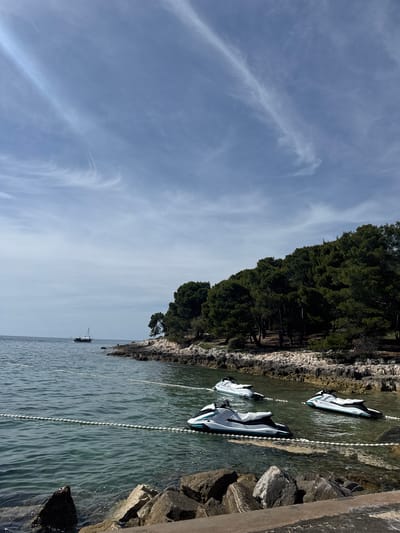 The image depicts a coastal scene in Općina Funtana, Croatia. The sky is a pale blue with wispy cirrus clouds. The coastline features a rocky shore with dense evergreen trees. Three white and black Yamaha jet skis are tethered to the shore by white buoy chains. A small boat is visible in the distance on the water. Rocks and concrete appear on the right side of the image.