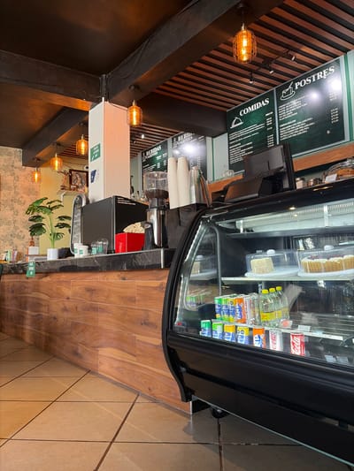 A cafe's interior shows a black-framed display fridge stocked with beverages and pastries. To its left, a menu board lists food and dessert options. A coffee machine sits atop a counter, next to a stack of disposable cups. A hanging light fixture adds a warm glow. The floor transitions from wood-look planks to tile, and a plant sits near the counter. The orientation of the image is rotated 90 degrees clockwise.