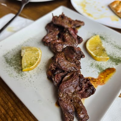 The image shows a close-up, eye-level view of a white rectangular plate holding a serving of grilled meat with two lemon wedges and some green herbs sprinkled on the plate. The meat appears to be thinly sliced and dark brown, with some glistening oil. One lemon wedge is on the left side of the plate, and another is on the upper right. The herbs are finely chopped and green, scattered around the meat and lemon. There is also a smear of orange sauce on the plate. The plate is set on a wooden surface, likely a table. In the background, parts of another white plate with some food remnants and a fork are visible. The overall setting suggests a restaurant in Atoluka, Bulgaria, where this dish is being served.