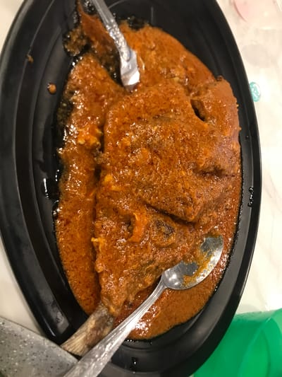 A plate of Asam Pedas fish dish on a black serving platter with a spoon and fork. The scene is Blangbitra, Indonesia. The fish is covered in a thick, reddish-brown sauce. The metal spoon and fork are partially submerged in the sauce. A green container is visible in the background.