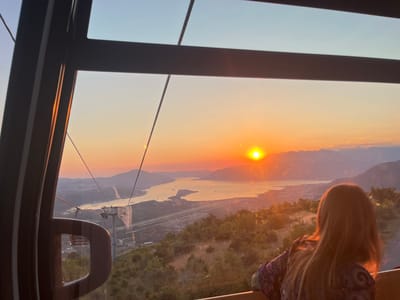 A young girl with long, light brown hair, wearing a purple and patterned shirt, is seen from behind, looking out of a cable car window at a scenic sunset over the Bay of Kotor near Čavori, Montenegro. The sun is a bright orb on the horizon, casting a warm, orange glow across the sky and water. Mountains frame the bay, and a town is visible along the coastline. The cable car interior, with its dark frame and cables, is partially visible in the foreground, suggesting the perspective is from within the moving vehicle. The overall scene conveys a sense of peace and awe at the natural beauty.