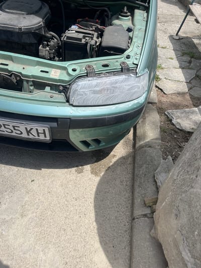 The image shows the front of a light green car, possibly a Fiat, with its hood open, revealing engine components such as the battery and fuse box. The car's license plate, partially visible, includes the characters "255 KH." The headlight has a cloudy or scratched appearance. The car is parked on a concrete surface, likely a sidewalk or driveway. The environment includes a stone wall or large rock formation in the background, suggesting the location could be in a residential area or near some natural features. Based on the location mentioned, the car is possibly in Slashten, Bulgaria.