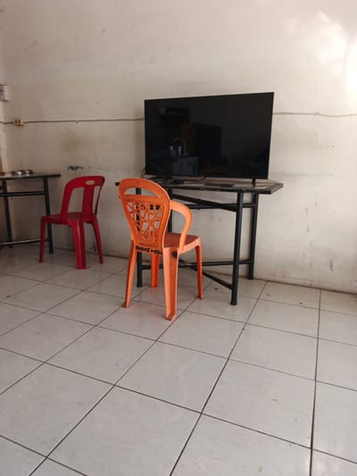 In Geudonggeudong, Indonesia, an indoor scene depicts a room with tiled flooring and plain walls. A large, black flat-screen television is mounted on a dark metal stand. In front of the stand, two plastic chairs are positioned: one is red, and the other is orange. To the left of the television stand, another dark metal table is visible, with a small round object, possibly an ashtray, on its surface. A single electrical outlet is visible on the wall to the left of the scene, with a thin wire running horizontally from it. The lighting appears to be natural, coming from the right side of the frame, casting shadows on the floor.