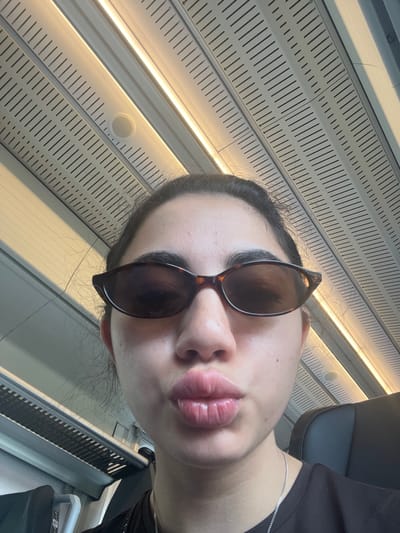 A young woman with dark hair and sunglasses is taking a selfie in what appears to be the interior of a train. Her lips are pursed for a kiss. The ceiling of the train car is visible, featuring fluorescent lighting and a perforated panel. The woman is wearing a black t-shirt and a silver necklace. The context of Vejprnice, Czechia, is not discernible from the visual information provided in the image.