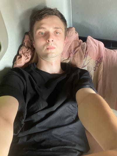 A young Caucasian man with light brown hair and green eyes is lying down. He is wearing a black t-shirt and is partially covered by a patterned blanket that appears to be pink and beige. The background suggests an indoor environment, possibly a vehicle or a room with a textured wall and a piece of furniture. The lighting is dim, casting shadows on the man's face and the surrounding objects. The man's gaze is directed towards the camera, and he has a neutral expression. There is no indication of the location being in Brachterbeek, Netherlands.