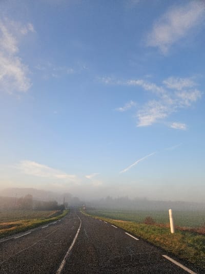 The scene depicts a rural road in Orcemont, France, under a light blue sky with scattered wispy white clouds and a faint contrail in the upper right quadrant. The lower half of the landscape is enveloped in dense fog or mist, obscuring distant features and the horizon. A two-lane asphalt road with a continuous white center line and dashed white shoulder lines curves gently into the fog towards the distant vanishing point. The road surface appears damp. On either side of the road, a strip of green grass and dirt borders the asphalt, leading to fields of green vegetation. Through the mist, trees are faintly discernible in the background on both sides. A white, cylindrical road marker stands upright on the right shoulder of the road in the mid-ground. No individuals, vehicles, or discernible activities are present in the image.