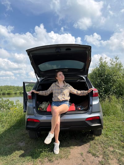 A young woman sits with her legs crossed in the open trunk of a grey SUV. She wears a tan and brown plaid shirt and denim shorts, with white sneakers. The car is parked on a dirt path next to a grassy area with green bushes and trees. A body of water, likely a lake, is visible in the background to the left, with a line of trees on the opposite bank. The sky is blue with scattered white clouds. The location is Gushchino, Russia.