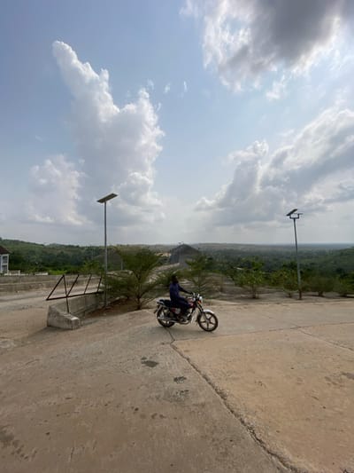 The scene depicts an elevated, concrete paved area in Ebuwana, Nigeria, under a bright sky with scattered cumulus clouds. A single adult male, dressed in dark blue clothing, sits astride a silver and red motorcycle with chrome accents, positioned centrally on the concrete surface. He faces forward and slightly to the right. The motorcycle is stationary or moving slowly. To the left of the motorcycle, a concrete retaining wall with a dark metal railing is present. Two tall, grey metal streetlights, each topped with a dark rectangular solar panel and a light fixture, are visible; one to the left of the motorcycle and one further right. Small, green-leafed shrubs are planted intermittently along the concrete edges. The background features an expansive, undulating landscape of dense green foliage, indicating hills or forested terrain. A large, skeletal metal-framed structure with a light-colored roof is discernible on the distant horizon. A portion of a light-colored building with a dark roof is visible on the far left edge of the frame. The concrete ground exhibits visible cracks and minor discoloration. The scene is illuminated by strong daylight, casting soft shadows.