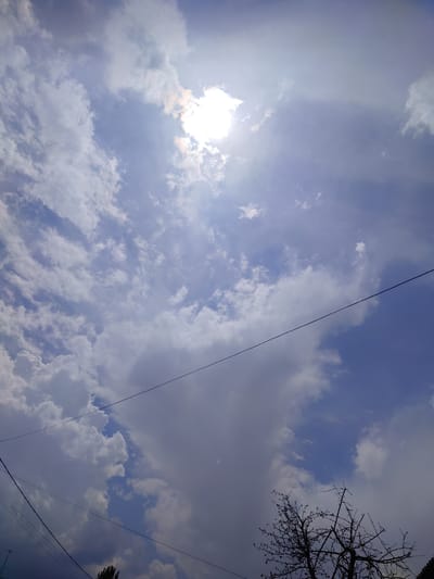 This is a low-angle, vertical shot of a sky filled with clouds and the sun. The sun is a bright, hazy orb in the upper portion of the image, partially obscured by wispy, white clouds. Larger, darker clouds are present in the lower half, casting shadows and creating a contrast with the brighter sky. Several thin, dark wires are visible diagonally across the frame, suggesting the presence of utility lines. In the bottom right corner, the silhouetted branches of a deciduous tree are visible against the sky. The overall impression is of a daytime scene with a mix of sun and clouds, possibly indicating changeable weather. The location context of Кашка-Суу, Kyrgyzstan, suggests this is an outdoor scene in that region. There is no visible text in the image.