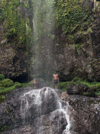 A vertical shot shows a shirtless man in dark-colored shorts standing behind a waterfall. His body is tan, and he is facing the camera. The waterfall is white and cascades down dark gray rocks. On either side of the waterfall are steep, rocky cliffs covered in green moss and vegetation. The location is in the city of Budar, Nepal.
