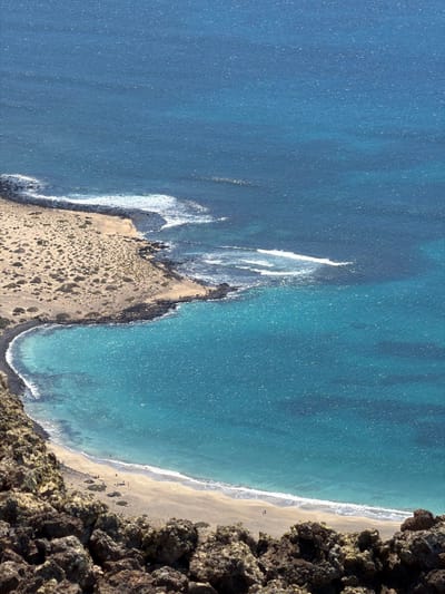 The image shows an aerial view of a coastline in Haría, Spain. A sandy beach stretches along the edge, meeting the turquoise ocean. Waves break along the shore, creating white foam. Rocky cliffs frame the right side of the image, with a sandy peninsula jutting out into the water on the left. Scattered vegetation covers the peninsula. Several people are visible on the beach.