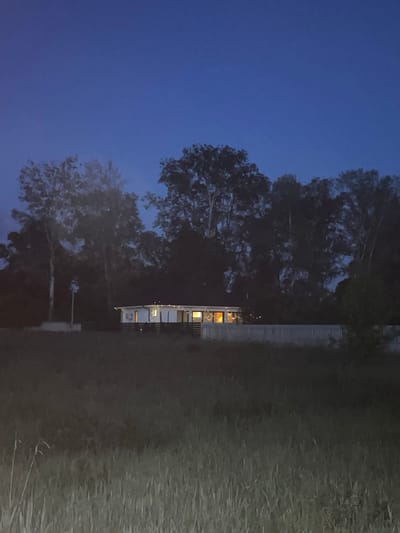 This image depicts a static nocturnal scene in Petrivka, Ukraine. The upper half of the frame is dominated by a clear, deep blue sky. A dense line of tall, dark-foliaged trees forms a backdrop across the middle ground, extending to both sides of the frame. Centrally positioned in the middle ground is a single-story house featuring a dark roof and white exterior walls with dark vertical elements. The house is illuminated from within, with multiple windows emitting a warm yellow-orange glow. Small, round, white exterior lights are visible on its facade. To the immediate left of the house, an upright pole with a white rectangular object is situated amidst the trees. A light-colored, horizontal slatted fence runs in front of the house, separating it from the foreground. The foreground consists of an expanse of tall, unkempt grass, rendered in shades of dark green and grey under low ambient light. No individuals or active subjects are present.