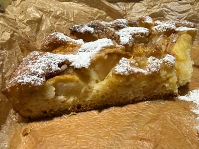 A close-up, overhead shot shows a piece of apple cake dusted with powdered sugar, placed on crinkled brown parchment paper. The cake appears to be baked in a rectangular pan, and it has a golden-brown crust with visible chunks of apple throughout. The parchment paper is also golden-brown and textured, with some shiny spots, possibly from oil or butter. The lighting is warm and casts soft shadows. There are no people or other objects visible in the image. The scene appears to be a close-up of food preparation or presentation. The location in Simonikha, Russia, is not discernible from the image.