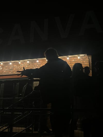 A dark, low-angle shot taken at night, possibly in Dundee, United Kingdom. A brick wall is lined with small, round lights, possibly string lights. A figure, silhouetted against the wall, appears to be reaching out with a hand. A faint inscription "AVANNA" is visible in large, stylized letters, angled and partially obscured in the background. Metal bars are visible on the right, possibly from a railing or structure.