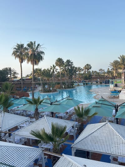 The image depicts an outdoor resort area in Taguermess, Tunisia, under a clear, light blue sky. The central feature is a large, irregularly shaped swimming pool with bright blue water, containing a small, circular island adorned with a palm tree and blue mosaic tiling. Several individuals are visible in and around the pool; one person appears to be swimming, while others are standing or lounging on the paved deck.
Numerous tall palm trees are interspersed throughout the entire scene, framing the pool and extending into the background, where more lush vegetation and distant structures are faintly visible. Along the poolside, several straw-roofed palapas provide shade, and blue sun loungers are arranged on the paved areas. To the right of the pool, a temporary stage-like structure with speakers, a blue base, and green and white panels is present. A yellow-domed structure is visible among the trees in the middle distance to the left.
In the foreground, an extensive outdoor dining area is arranged. It features multiple white, slatted pergolas with pyramidal roofs, as well as several large, light teal sun umbrellas. Beneath these structures, numerous tables are meticulously set with white tablecloths, white chairs, and tableware, indicating readiness for service. Smaller palm trees grow throughout this dining area. The ground beneath the dining tables consists of a patterned surface of blue and terracotta-colored paving. No active dining or significant interaction is occurring in the immediate foreground.