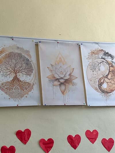 The image displays three artworks pinned to a light yellow wall, likely in an indoor setting. The artworks are arranged horizontally and feature a nature-inspired, spiritual theme. The leftmost artwork depicts a circular design with a stylized tree of life, its roots intricately intertwined. The central artwork showcases a delicate lotus flower with watercolor-like drizzles. The rightmost artwork presents a yin and yang symbol formed by two trees. Below these artworks, on the same wall, several red paper heart cutouts are affixed. The overall scene suggests a decorative or contemplative space, possibly within a home, studio, or communal area. There is no visible indication of Slashten, Bulgaria within the image itself.