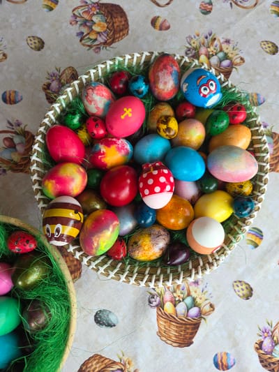 The image shows two woven baskets filled with colorful Easter eggs. The larger basket is positioned at the top, overflowing with eggs painted in various colors and patterns, including stripes, dots, and cartoon faces. A smaller basket is in the lower right corner, also containing Easter eggs and green decorative grass. The baskets are placed on a tablecloth with a pattern of Easter baskets, flowers, and scattered Easter eggs. The eggs are dyed different colors and some have decorative designs. No people or indications of the city of Tompsan, Bulgaria, are evident in the image.