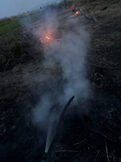 In Golyam Chardak, Bulgaria, at dusk, a small outdoor fire burns amidst dry vegetation and scattered branches. Thick white smoke billows from the scene, obscuring parts of the ground. A person in a pink shirt is visible in the background, seated near the fire. In the foreground, a green hose nozzle is spraying water onto the smoking embers, suggesting an attempt to extinguish the fire. The ground is covered in dark soil and dry, brown grass.
