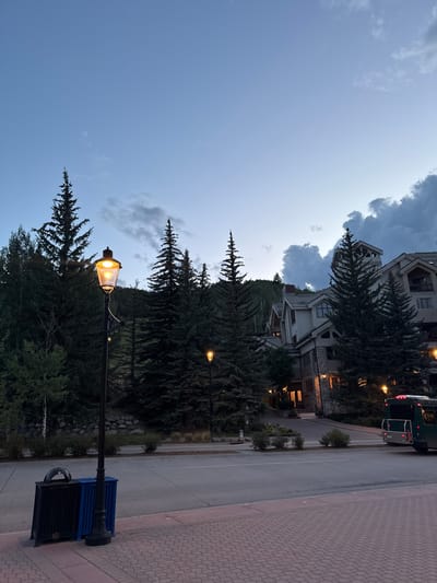 This image depicts a street scene in Beaver Creek, Colorado, USA, during twilight. A vintage-style lamppost with a glowing yellow bulb stands prominently in the foreground, to the left of the frame. Adjacent to the lamppost is a black trash receptacle and a blue recycling bin. The street is paved with red brick pavers in the foreground, transitioning to asphalt further back. On the right side of the street, a dark green shuttle bus with visible taillights is parked or moving away from the camera.

Behind the street, a multi-story building with a rustic architectural style, characterized by stone and wood accents, is visible. Numerous evergreen trees, likely pine or spruce, are scattered throughout the scene, with some forming a dense forest on a hillside behind the building. The sky is a gradient of deep blue at the top, transitioning to lighter shades near the horizon, with scattered clouds, some appearing dark and dramatic. The overall ambiance suggests a serene mountain resort town as evening sets in. No people are discernible in the image.