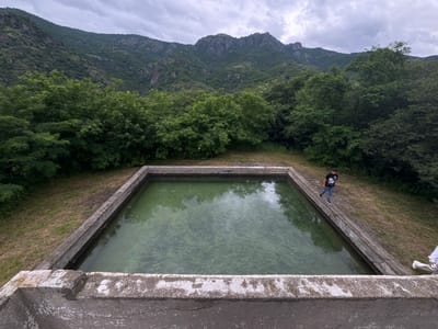 An aerial view shows a rectangular concrete reservoir filled with green water, located in a mountainous, forested area. The reservoir's edges are weathered concrete. A young male wearing a black t-shirt with white lettering and blue jeans is walking along the edge of the reservoir on the right side of the frame. Further to the right, a person in white pants is partially visible at the edge of the reservoir. The background is dominated by a steep, densely wooded mountain slope under a cloudy sky. The overall scene appears to be a natural landscape with a man-made water structure.
