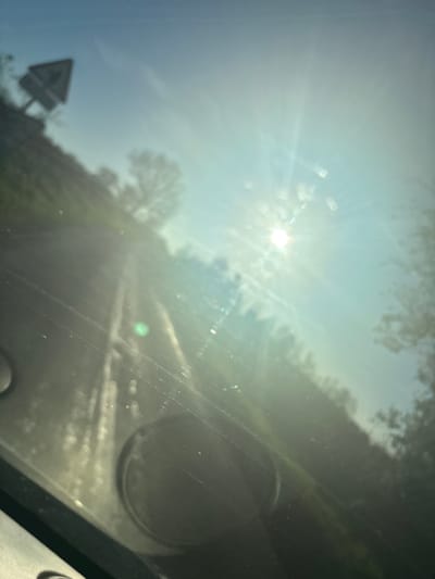 The image appears to be taken from inside a vehicle, looking through the windshield. The sun is shining brightly, causing significant glare and obscuring the view. A road is visible in the distance, flanked by trees. A road sign, triangular in shape, can be seen on the left side of the frame. The presence of a road sign suggests that the vehicle is located near or within Ruyton-XI-Towns, United Kingdom. The exact location cannot be determined without further details.