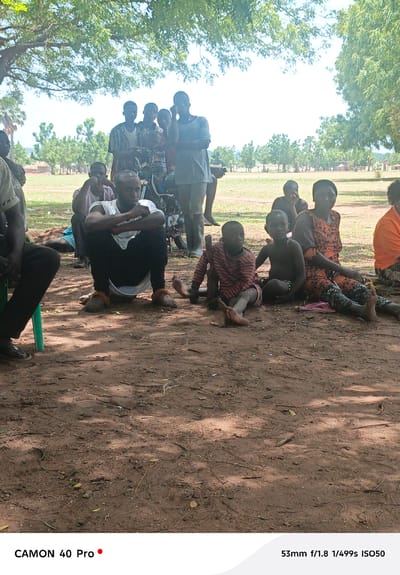 The image depicts a group of people gathered outdoors in Gidan Kogi, Nigeria. The ground is red soil, covered with scattered leaves and twigs, shaded by a tree. The group includes men, women, and children. One man is squatting in the foreground, wearing a white shirt and dark pants. A motorcycle is visible in the background, partially obscured by the standing figures. Some individuals are sitting on the ground, while others are standing. The environment appears rural, with trees and vegetation visible in the distance. The lighting suggests it is daytime.