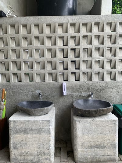The image displays an outdoor handwashing station, likely located in Kota Batu, Indonesia. The background features a grey concrete wall composed of two sections: an upper part with a grid of open, L-shaped breeze block voids, and a lower part with a rough, textured finish. Two individual washing units are positioned against the lower wall. Each unit consists of an irregularly shaped, dark grey, natural stone basin resting on a substantial, rough-textured, light-colored concrete or stone pedestal. A metal water tap is mounted on the wall directly above each basin. Centered between the two taps is a white, wall-mounted liquid dispenser. To the far left, a tall, dark-colored container holds several long-handled objects, possibly umbrellas, with yellow and brown handles visible. To the far right, the top edge of a green-lidded waste bin is partially visible. The ground surface is paved with light-colored bricks or pavers. In the upper background, behind the wall, a large black cylindrical object and white cables are discernible, with glimpses of green foliage. No individuals are present in the scene.