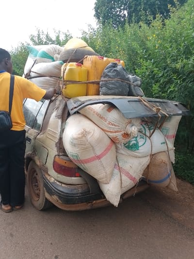 In Ugwolawo, Nigeria, a heavily overloaded, older model sedan, possibly silver or light-colored, is stationary on a paved road. The vehicle shows significant rust on its body panels, particularly around the rear bumper and wheel arches. Its roof and rear trunk area are completely covered with a precarious arrangement of cargo. This load includes multiple large white woven sacks, several yellow plastic jerrycans, a black sack, and what appears to be a spare tire. One visible sack is labeled "SUPERB FLOUR." The entire load is secured with a network of ropes. A person, seen from the waist up and facing away from the camera, is standing on the left side of the vehicle. They are wearing a yellow t-shirt and dark trousers, with a dark shoulder bag, and appear to be interacting with the ropes securing the cargo on the vehicle's roof. The immediate environment features a background of dense green foliage, consistent with tropical vegetation, under an overcast sky.