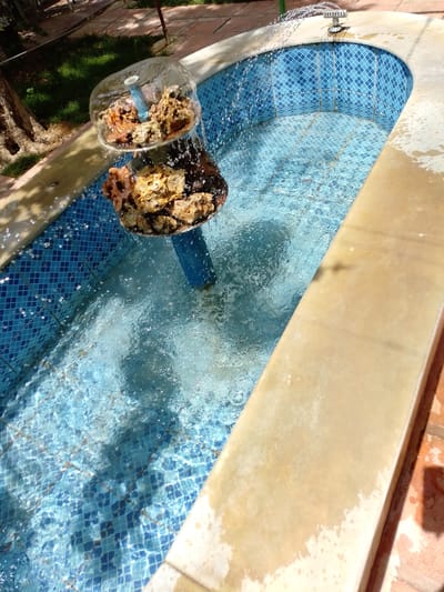 This image captures an outdoor fountain and pool area, likely located in a garden or courtyard in Qudsaiya, Syria, on a sunny day.

The main subject is a two-tiered fountain structure situated within a rectangular pool with a rounded end. The fountain's tiers are composed of clear, bowl-like containers filled with natural rock formations. A white pipe is visible in the upper tier, and a blue pipe supports the lower tier from the pool bed. Water actively cascades and splashes from these tiers into the pool, creating a dynamic surface. At the rounded far end of the pool, a metallic sprinkler head sprays arcs of water into the basin.

The pool is lined with distinct blue and light blue mosaic tiles arranged in a diamond pattern, visible through the clear, rippling water. A light beige or cream-colored coping surrounds the edge of the pool. Beyond the coping, parts of the surrounding environment are visible: green grass, the trunk of a tree, and terracotta-colored paving stones. The bright sunlight illuminates the scene, causing the water to sparkle and casting subtle shadows. No people are present, and there is no visible text in the image.