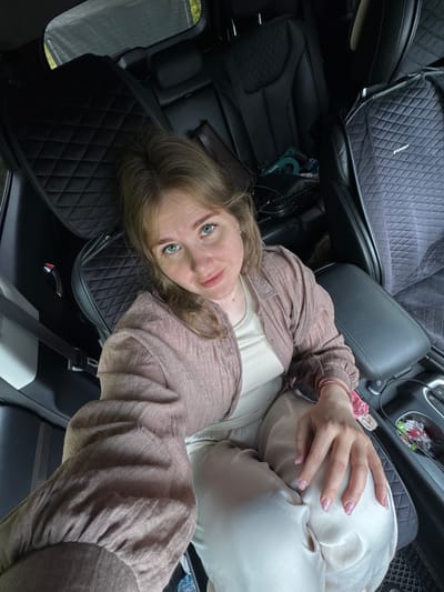 A woman with blonde hair and blue eyes is sitting in the back seat of a car. She is wearing a light-colored t-shirt and a pinkish-brown cardigan. The car's interior appears to be black leather with quilted seat covers. In the background, there is a view of trees and foliage, suggesting an outdoor location, possibly on the outskirts of Большие Кусты, Russia. The woman is looking at the camera, and her hands are resting on her lap. The lighting is natural, and the image appears to be a selfie.