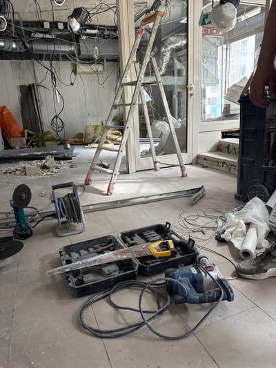 The image depicts an indoor construction site in Colombes, France, characterized by a chaotic arrangement of tools and debris. A silver A-frame ladder stands prominently, its legs wrapped in red tape. In the foreground, a turquoise Makita angle grinder with a blade sits on the floor, connected to a power cord. Next to it is a blue reel of electrical cable. A black tool case contains a jigsaw and a metal cutting blade. Further to the right, a blue and silver rotary hammer drill is positioned on the floor, its cord coiled. Several rolls of white plastic sheeting and other construction materials are scattered around. The ceiling is a tangle of wires, ventilation ducts, and exposed light fixtures, suggesting an ongoing renovation or demolition project. The overall environment is dusty and disorganized, with pieces of plaster and insulation visible on the floor.