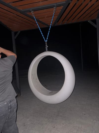 A person, seen from the chest down, is sitting on a paved surface next to a wooden and metal structure. They are wearing grey shorts and a grey t-shirt. The structure appears to be part of an outdoor playground or recreation area. Hanging from the structure by blue ropes is a large, white, ring-shaped object. The scene is dimly lit, suggesting it is nighttime or very early morning. The paved surface is made of small, square tiles. There is visible text on the metal structure, appearing to read "EGORLYKSKAYA".