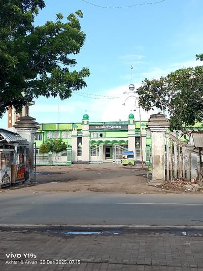 A daytime shot captures the Masjid Al-Muttaqien Jagamima, painted green and white. The mosque sits beyond a spacious courtyard, littered with debris, accessed through a gated entrance. The foreground shows a paved road, with structures on either side of the gateway partially obscured by trees and utility poles. A street vendor's cart is stationed in front of the mosque. The photo's lower left corner displays a vendor's stall and the text "vivo Y19s" with the names "Akhtar & Arvan" and date "20 Des 2025, 07.15".