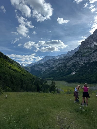 Two young women are standing in a grassy meadow with rolling hills and mountains in the background. One woman is wearing a white tank top, a red and black plaid shirt tied around her waist, and white socks. The other woman is wearing a pink tank top and a black skirt. They are both looking away from the camera, towards the mountains. The meadow is covered in green grass and scattered with small yellow flowers. The mountains are tall and rugged, with some patches of snow on their peaks. The sky is blue with white clouds. The scene appears to be a remote, natural landscape, possibly in the Alps of Albania.