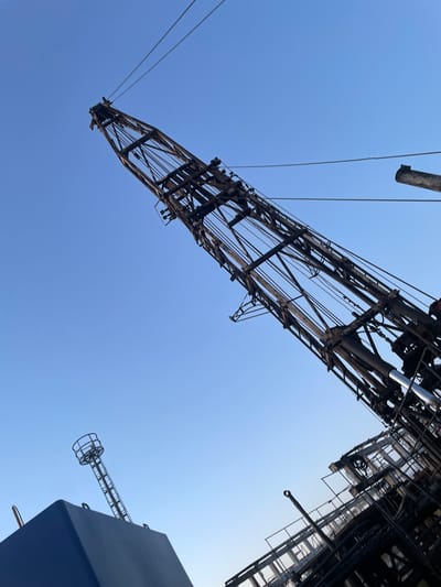 The image depicts a low-angle view of a large, rusty crane structure against a clear blue sky. The crane's boom, made of metal struts and cables, extends diagonally upwards from the lower right to the upper left of the frame. In the lower left corner, a portion of a large, dark blue, angular structure is visible, possibly a part of a ship or industrial equipment. A tall, thin metal tower with a circular cage-like structure at its top rises from this blue element, also angled against the sky. The overall scene suggests an industrial or maritime setting, possibly a port or shipyard. The location is identified as Michayol', Russia, though this specific context is not visually evident from the image itself. There are no people or any discernible actions taking place in the photograph. The environment is characterized by clear, bright daylight.