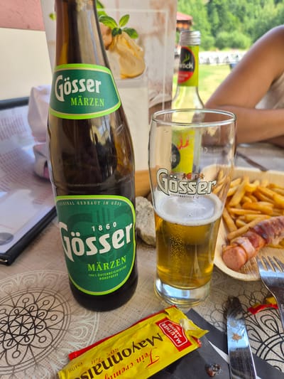 The image depicts a close-up view of a table setting, likely outdoors, with a focus on food and beverages. A tall, dark brown glass bottle of Gösser Märzen beer stands to the left, its green label prominent. Next to it, a beer glass filled with Gösser Märzen beer shows a frothy head and bubbles. In the background, a blurry bottle of Hirschwasser and a person's arm and shoulder are visible. To the right of the beer glass, a wooden plate holds a portion of French fries and a sausage wrapped in bacon. A packet of Mautner Markhof Feine Mayonnaise is situated in the foreground, alongside a knife and fork. The table is covered with a patterned tablecloth. The setting appears to be in Landl, Austria, suggesting a casual dining or picnic environment.