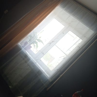 The image depicts an interior scene looking out a window, likely in Balakhna, Russia. The viewpoint is from within a room with dark walls and a diagonally oriented orange curtain partially covering the left side of the frame. A sheer white curtain is drawn across the window, diffusing the bright daylight outside. The window itself is a multi-pane casement style, with the frames appearing white. Through the translucent curtain and the window, one can discern the faint shapes of foliage, suggesting an outdoor environment with trees or bushes. A potted plant with green leaves is visible on the windowsill, and the pot is a muted purple. A portion of the room's interior wall, possibly a radiator or heating element, is visible near the bottom left, covered in a textured material. In the bottom right corner, a red-capped bottle, possibly a beverage, is partially visible against the dark wall. The overall lighting is characterized by the bright glare from the window, with the interior of the room being relatively dark.