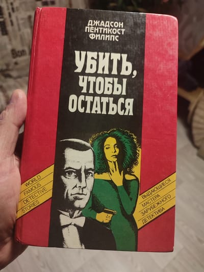 A hand holds a hardcover book with a red cover and a black spine. The title on the spine is in Russian and reads "УБИТЬ, ЧТОБЫ ОСТАТЬСЯ", which translates to "KILL TO REMAIN". Above the title are the authors' names: "ДЖАДСОН ПЕНТИКОСТ ФИЛИПС". On the left side of the book cover, a yellow banner contains the text "WORLD FAMOUS DETECTIVE STORIES" in English, also printed vertically. On the right side of the cover, another yellow banner contains Russian text that reads "ВЫДАЮЩИЕСЯ МАСТЕРА ЗАРУБЕЖНОГО ДЕТЕКТИВА", translating to "OUTSTANDING MASTERS OF FOREIGN DETECTIVE". The cover features a black and white illustration of a man and a woman. The man is in the foreground, looking towards the viewer, wearing a suit and bow tie. The woman is depicted in a green dress, holding a pistol, with her hand near her face. The background of the illustration is dark. The book appears to be in good condition, with some minor wear on the edges. The image is taken indoors, with a blurred background that suggests a domestic setting. No information about the city of Большое Афанасово, Russia is present in the image.